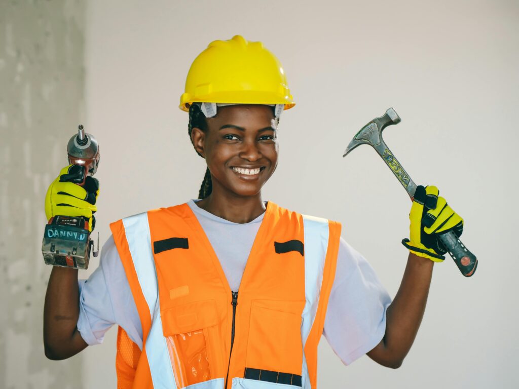 Smiling female engineer wearing safety gear, holding a drill and hammer confidently.